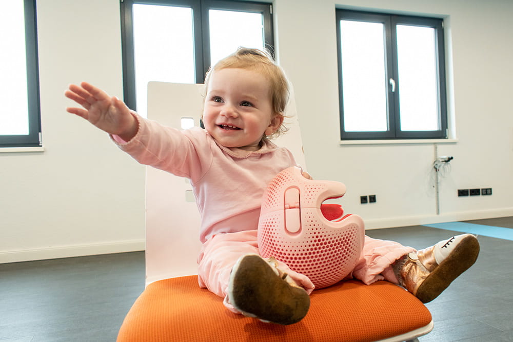 Ein Kleinkind sitzt auf einem Stuhl und hält eine Talee Kopforthese in der Hand