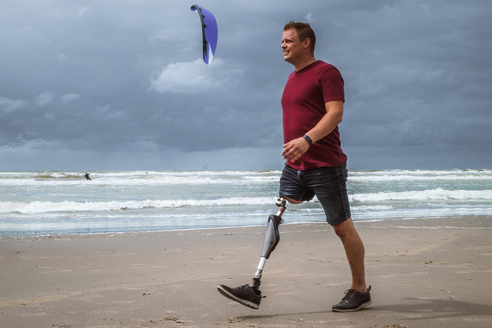 Ein Mann mit Endo-Exo-Prothese läuft am Strand entlang und hinter ihm ist ein Kitesurfer im Wasser.