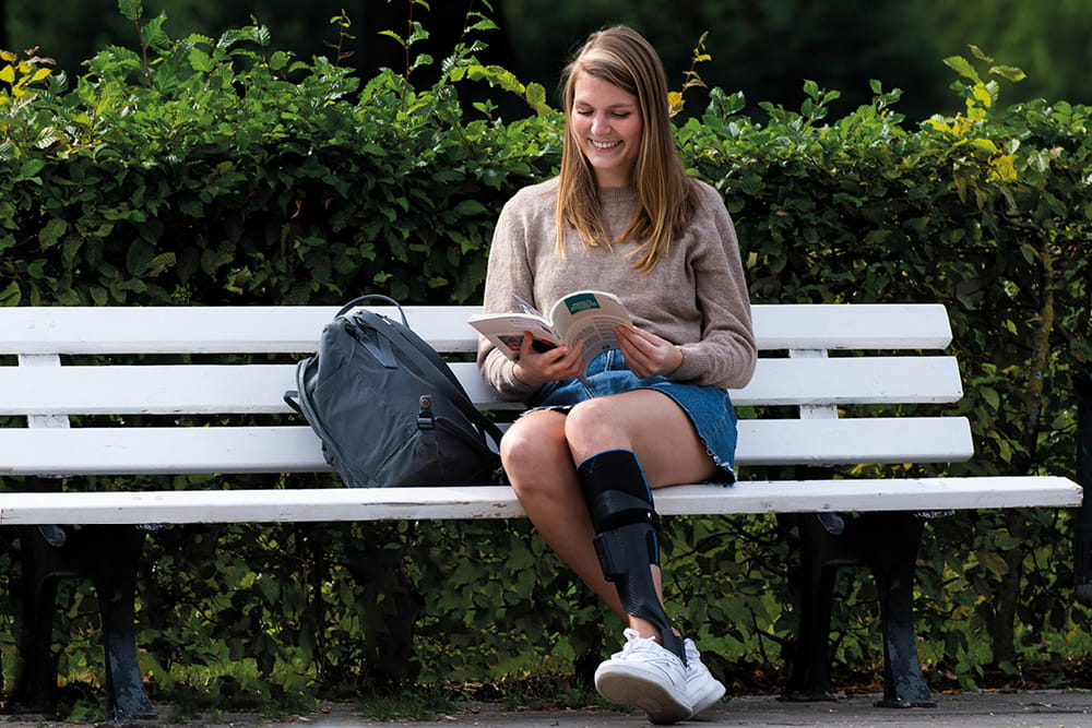 Eine Frau mit Beinorthese sitzt auf einer weißen Bank und ließt in einem Magazin.