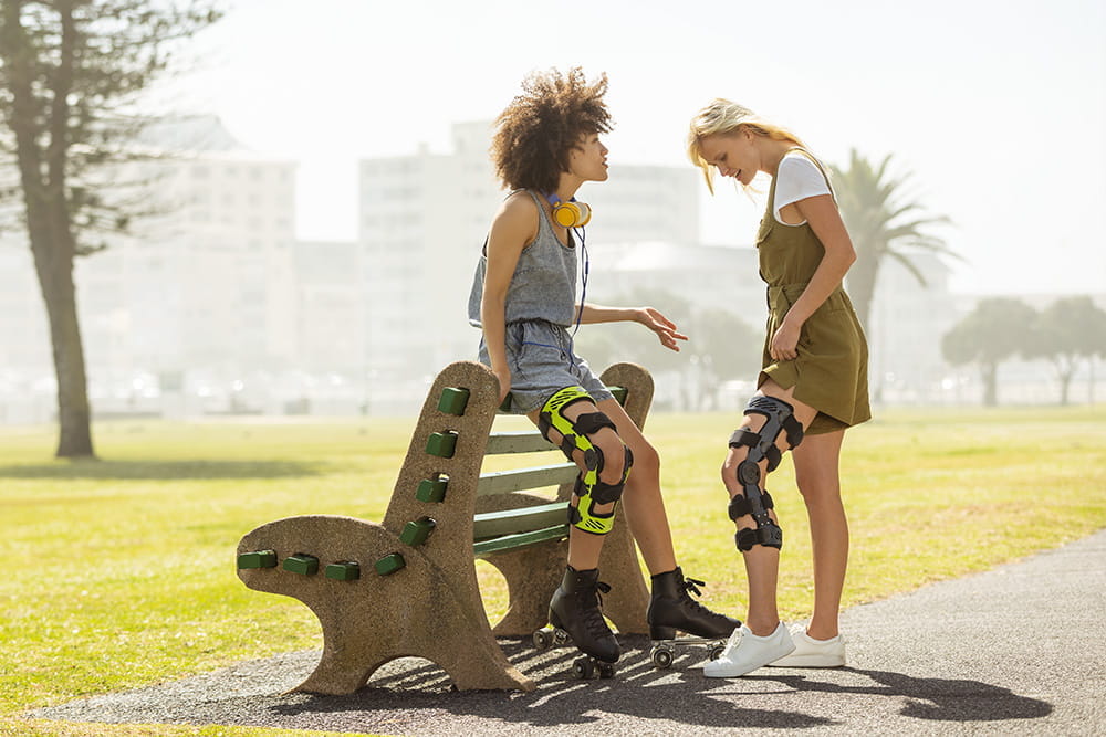 Zwei Frauen stehen an einer Bank und unterhalten sich. Beide tragen eine Orthese von Bauerfeind am Bein.