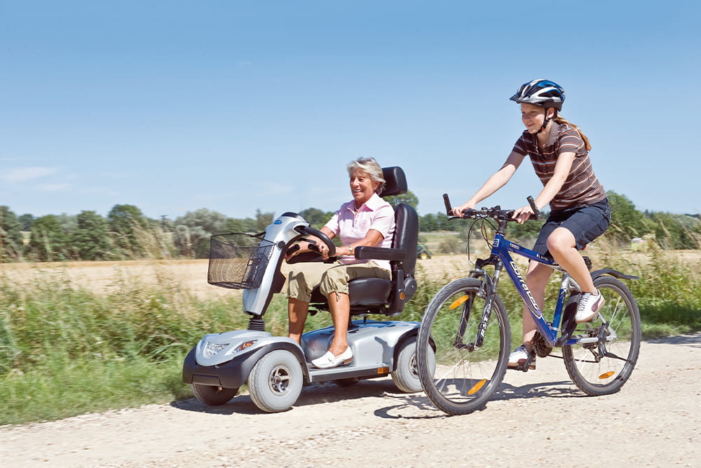 Eine Frau mit E-Scooter und eine Frau auf dem Rad fahren gemeinsam einen Sandweg entlang.