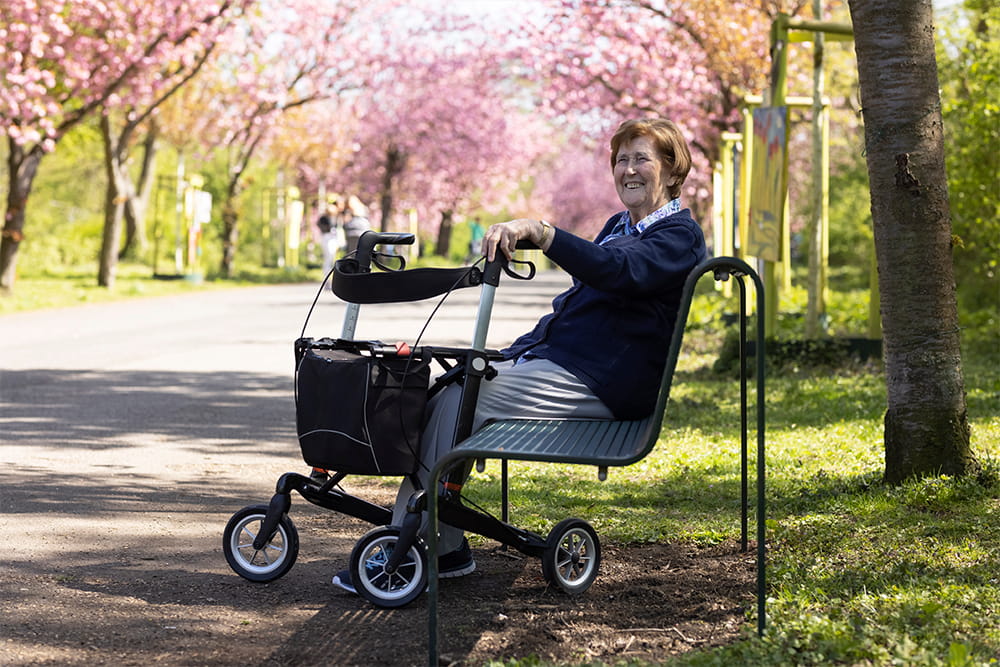 Eine Frau mit Rollator sitzt auf einer Parkbank.