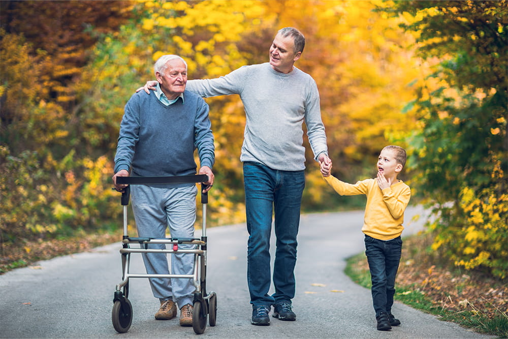 Eine Familie bestehend aus Opa, Vater und Sohn laufen einen Weg im Herbst entlang. Der Opa nutzt einen Rollator.