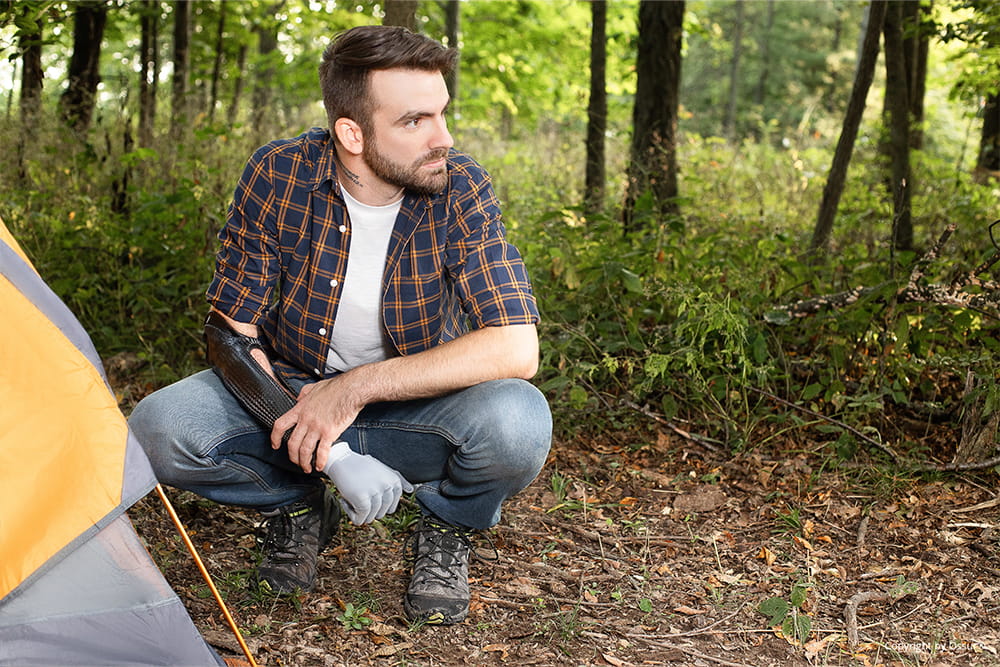 Ein Mann mit Armprothese hockt neben seinem Zelt im Wald.