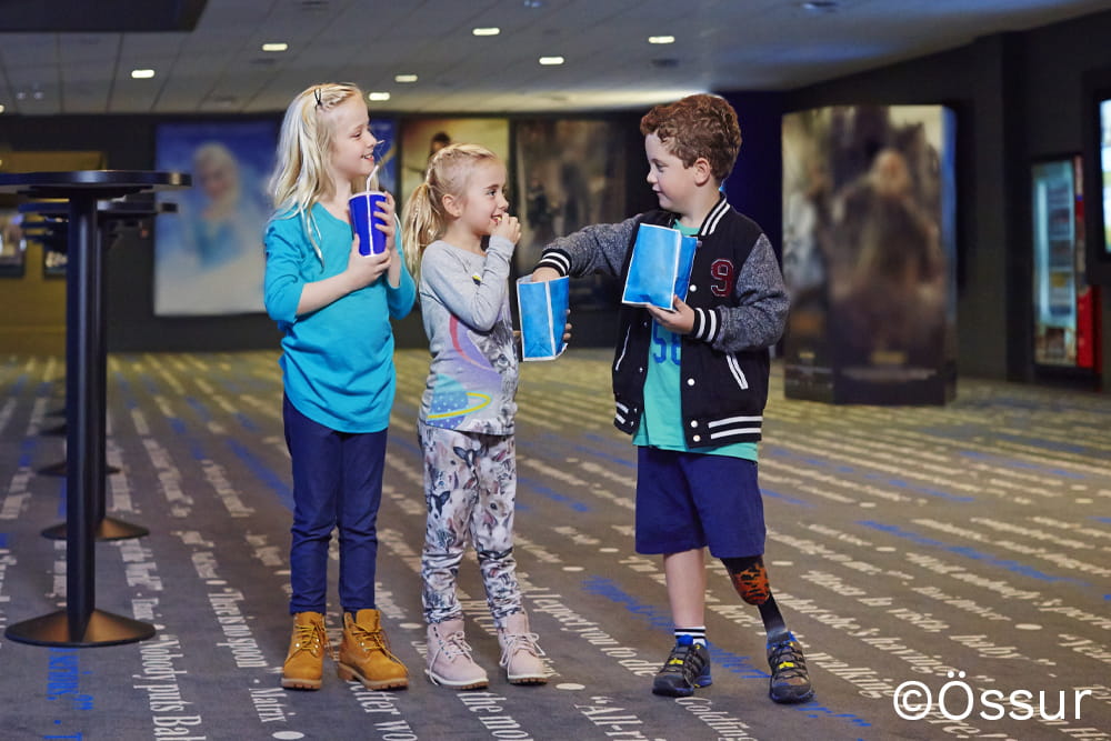 Drei Kinder stehen im Kinovorraum zusammen und essen Popcorn. Eines der Kinder trägt eine Beinprothese.