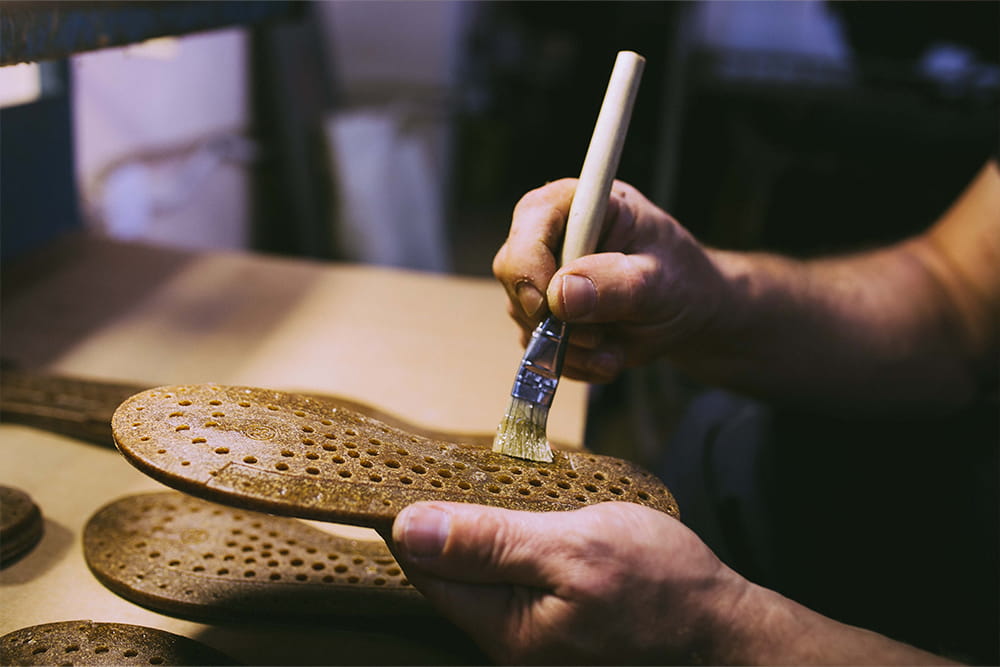 Eine Person hält eine Sohle in der Hand und streicht mit einem Pinsel einen Klebestoff darauf breit.