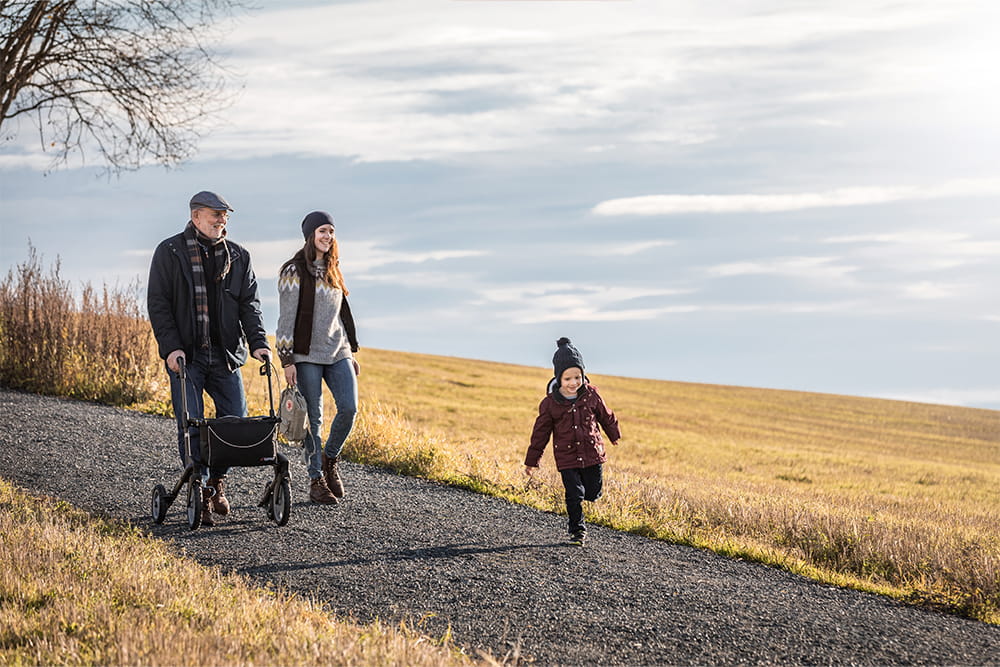 Ein Opa mit Rollator geht mit seiner Tochter und dem Enkel spazieren auf einem Feldweg.