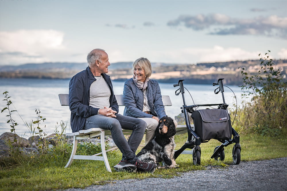 Zwei ältere Personen sitzen auf einer Bank in der Natur, neben der Bank steht ein Rollator von Topro.